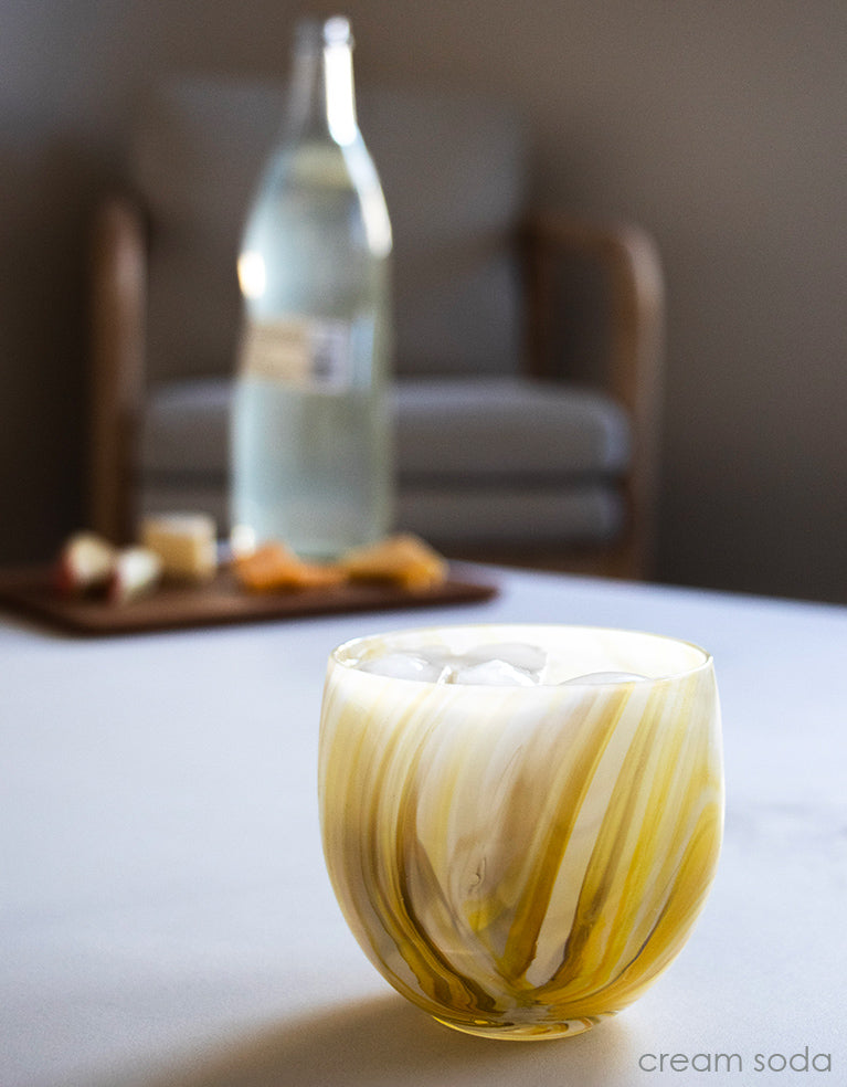 cream soda, yellow swirled hand-blown drinking glass on a white surface with a tray of orange slices and a bottle in the blurred background.