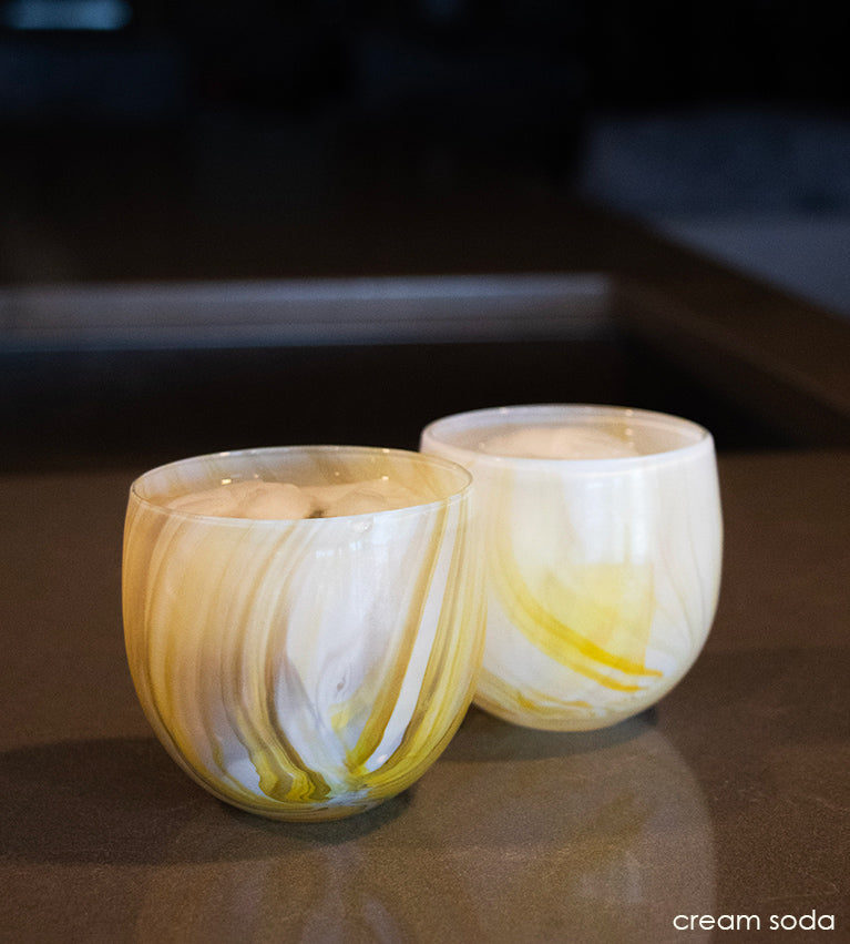 two cream soda, yellow swirled hand-blown drinking glasses on a brown marble counter. both glasses filled with ice and liquid.