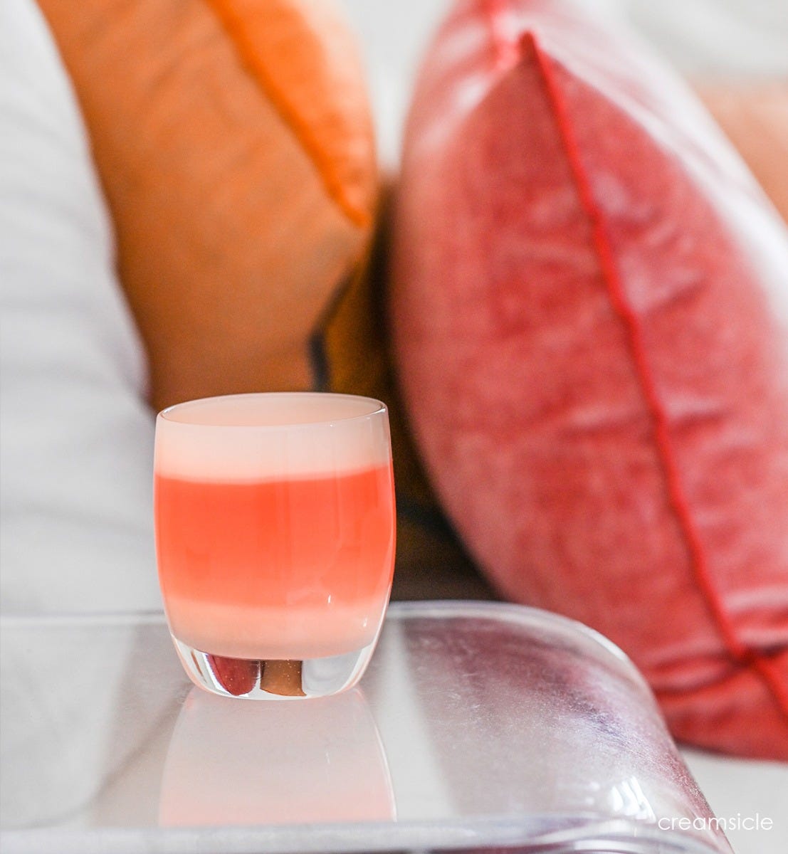 creamsicle orange creamed hand-blown glass votive candle holder on a clear living room table with orange and red throw pillows in background.