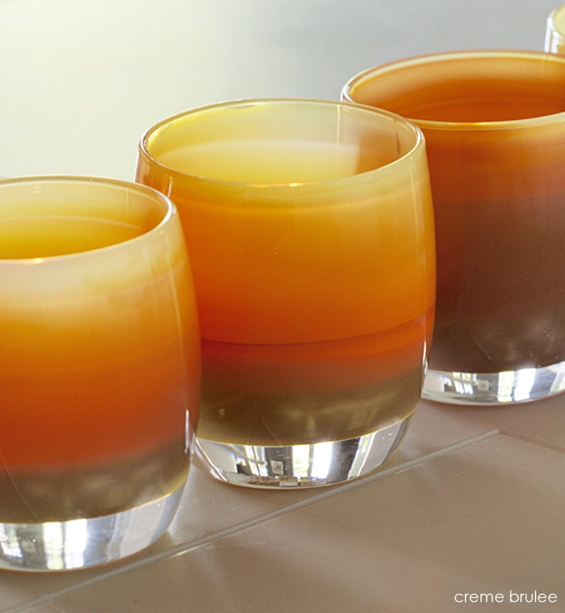 three creme brûlée caramel, hand-blown glass votive candle holders in a row on a tiled surface.