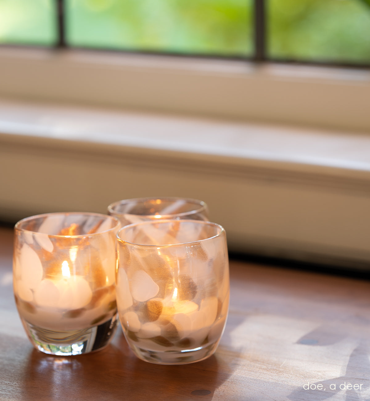 three doe, a deer shimmering gold and white petal, hand-blown glass votive candle holders on a wood surface in front of a white and metal framed window.