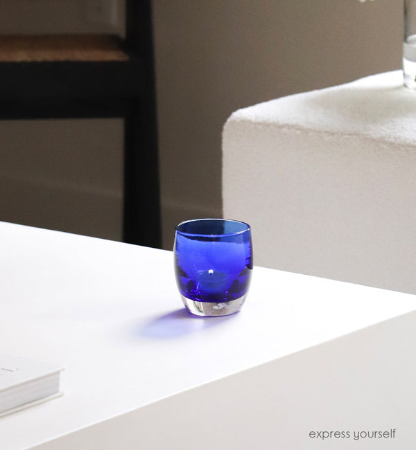 express yourself, transparent admiral blue, hand-blown glass votive candle holder on a white coffee table next to a plush white couch.