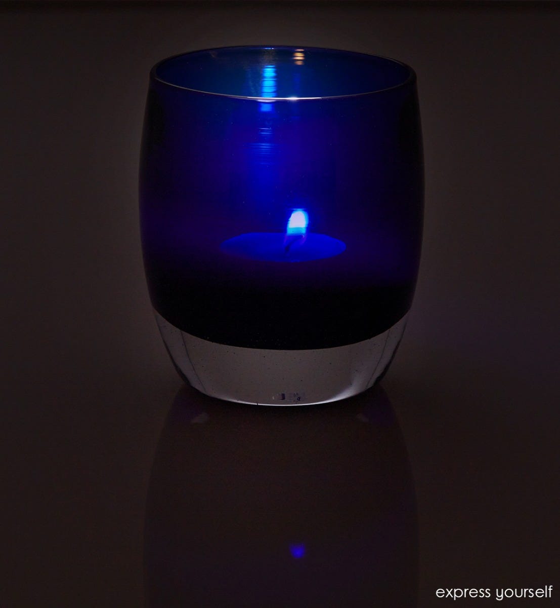 express yourself, transparent admiral blue, hand-blown glass votive candle holder lit in a dark room against a reflective surface.