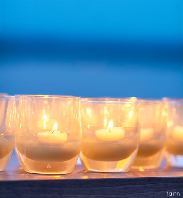 several faith translucent cream hand-blown glass votive candle holders lit at dusk on a wood table.