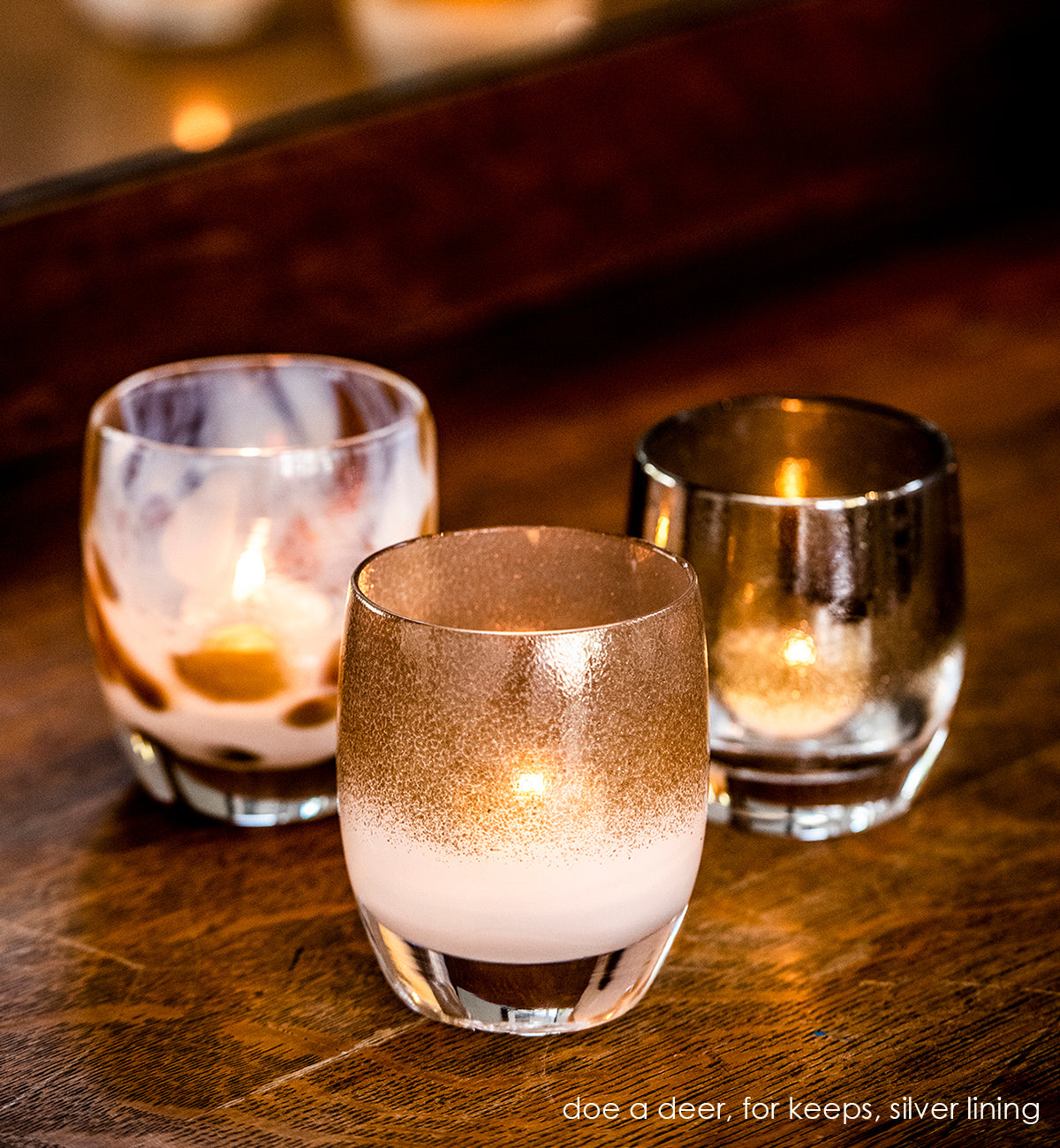 for keeps, half white half shimmering pewter hand-blown glass votive next to silver lining and doe, a deer glassybaby. the three are sitting lit on a dark wood table.
