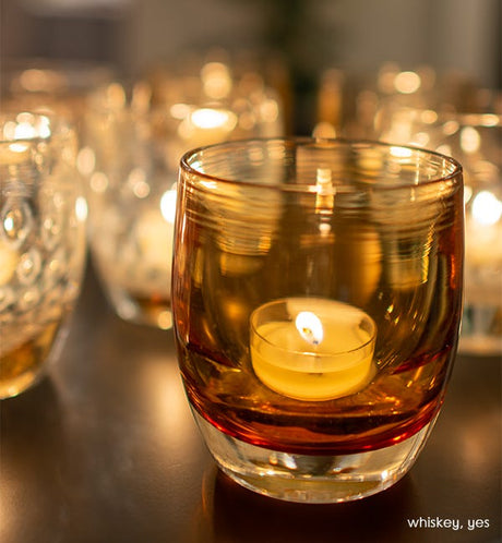 whiskey, transparent light amber, hand-blown glass votive candle holder. Paired with yes on a dark reflective surface.