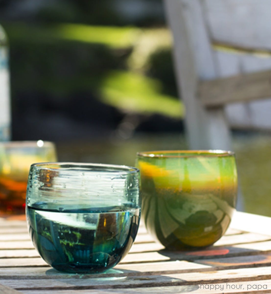 happy hour drinker, natilus blue hand-blown drinking glass with papa drinker on a wood patio table next to wood patio chair.