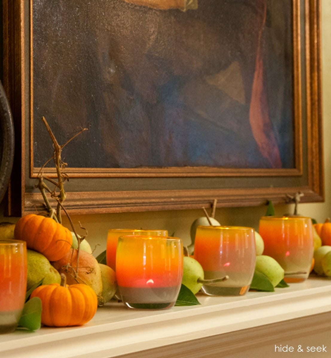several hide and seek autumn tone hand-blown glass votive candle holders on a mantle with decorative pumpkins and pears.