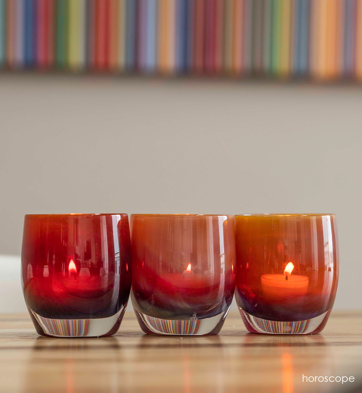 three horoscope purple orange, hand-blown glass votive candle holders on wood table with bookshelf behind.
