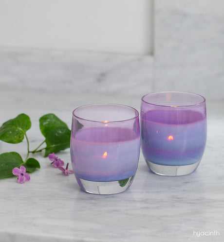 two hyacinth amethyst hand-blown glass votive candle holders on a marble countertop next to purple flowers.