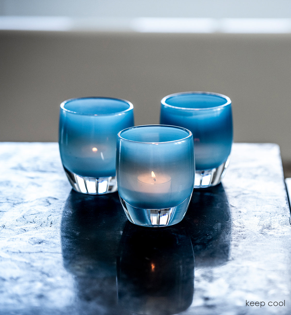 three keep cool light blue hand-blown glass votive candle holders with tealights lit on a dark marble countertop.