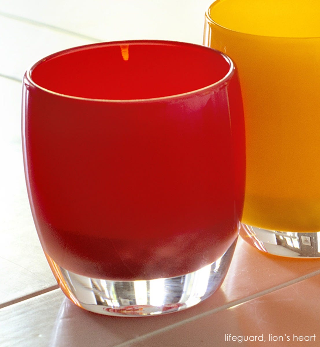 lifeguard hand-blown cherry red glass votive candle holder next to yellow lion's heart on cream table.