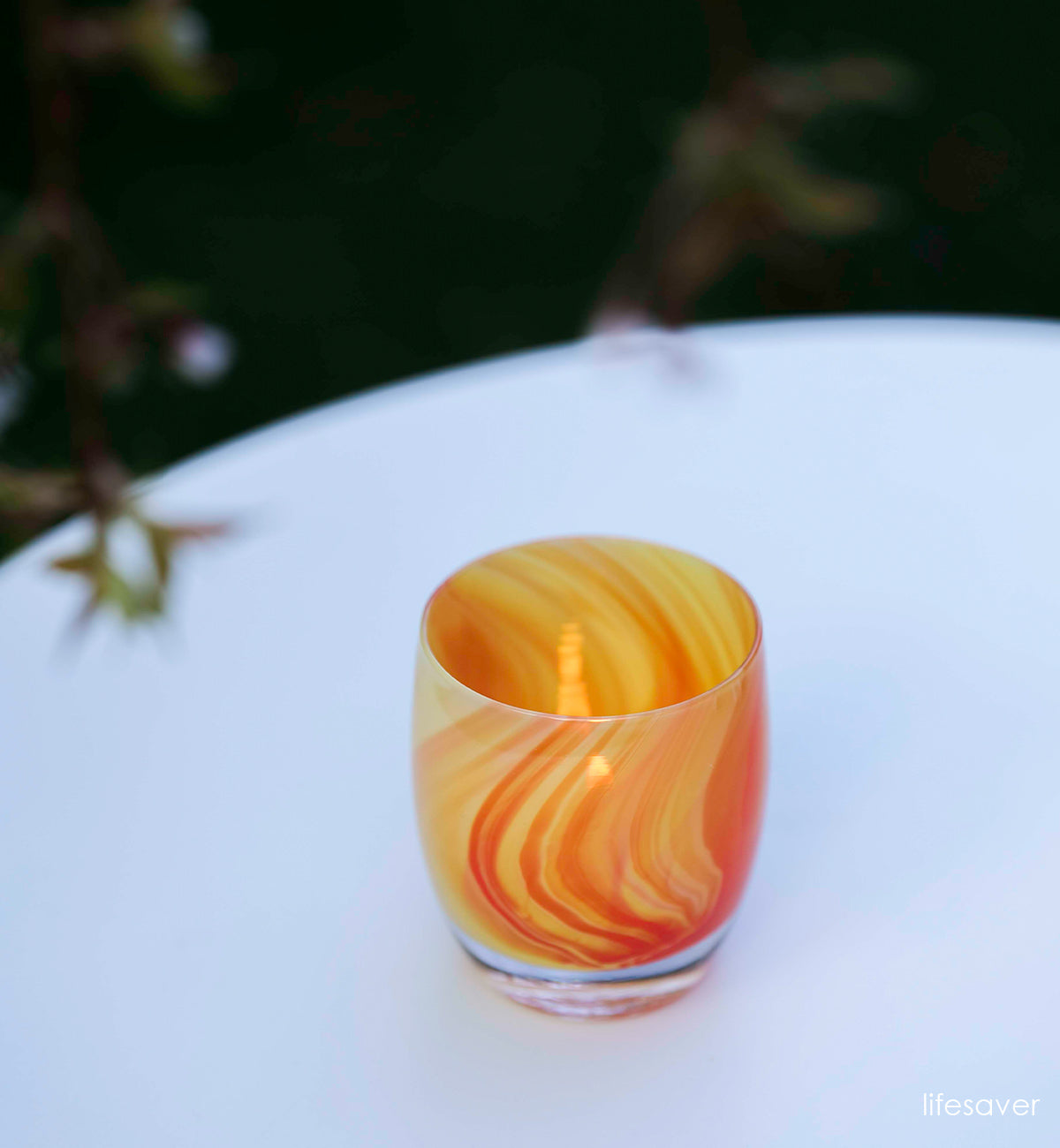 lifesaver orange and yellow swirled, hand-blown glass votive candle holder lit on a table outside with a white tablecloth.