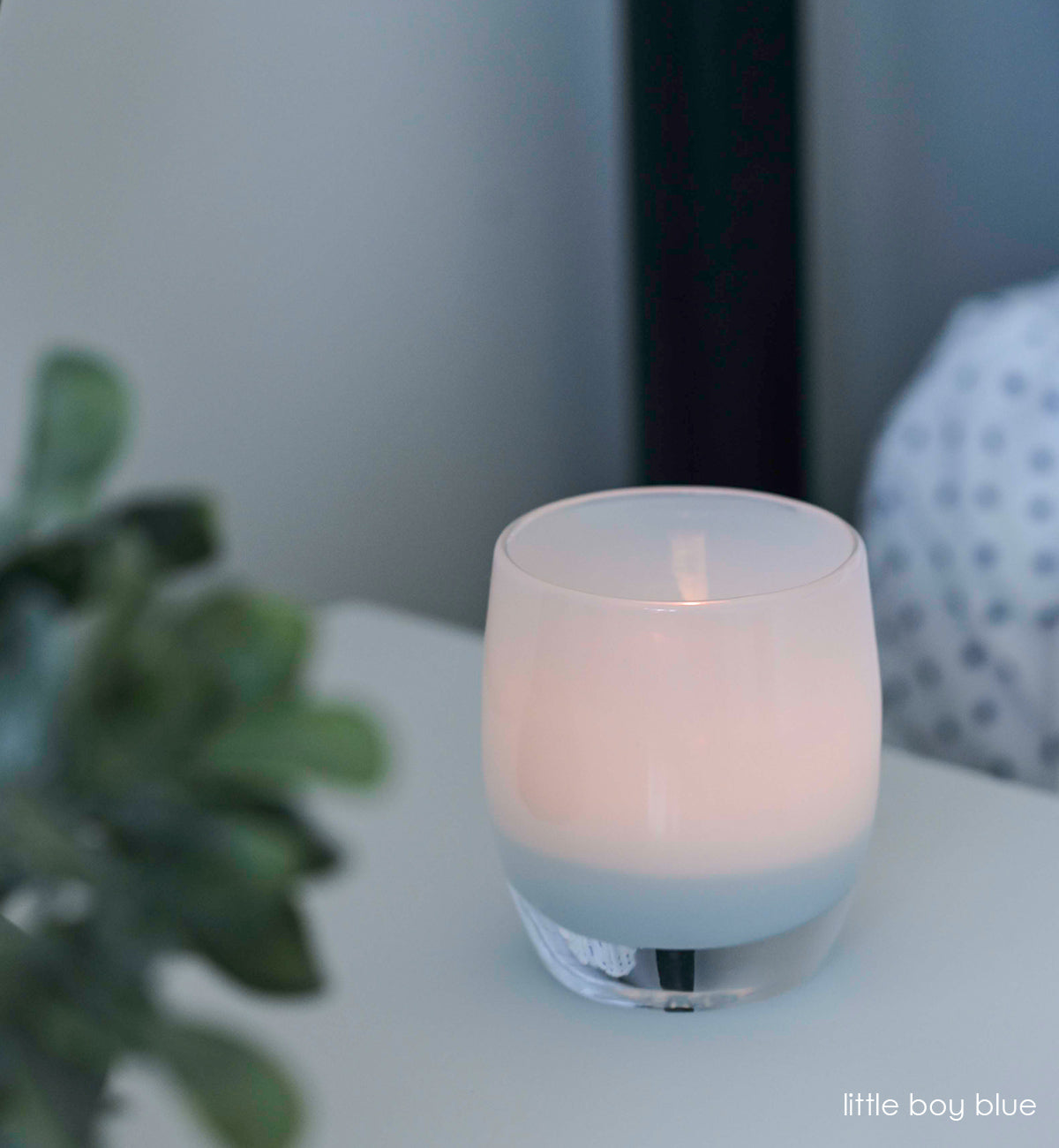 little boy blue, soft blue hand-blown glass votive candle holder on white table next to green plant.