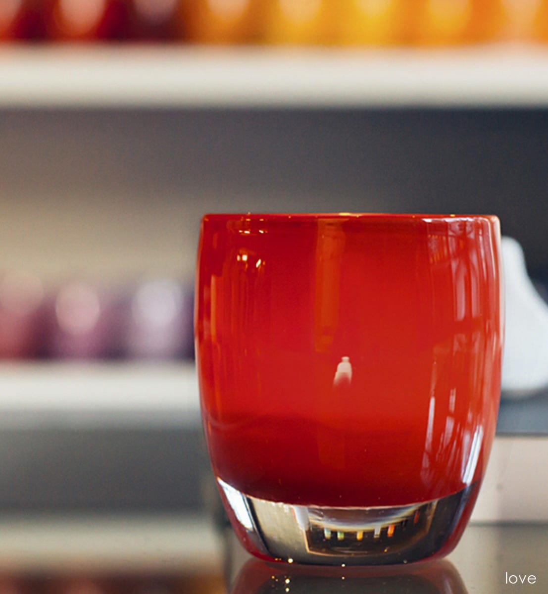 love brick red hand-blown glass votive candle holder with glassybaby shelves blurred in the background.
