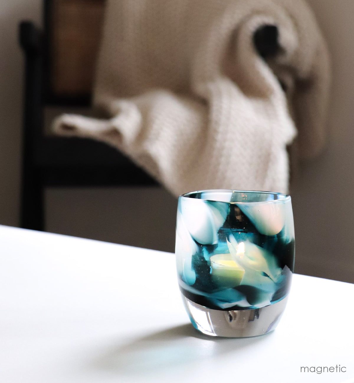 magnetic teal and white petal textured, hand-blown glass votive candle holder on a white table with a chair and blanket in background.