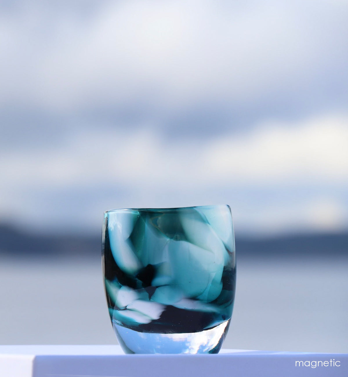 magnetic teal and white petal textured, hand-blown glass votive candle holder with mountains and lake blurred behind it.