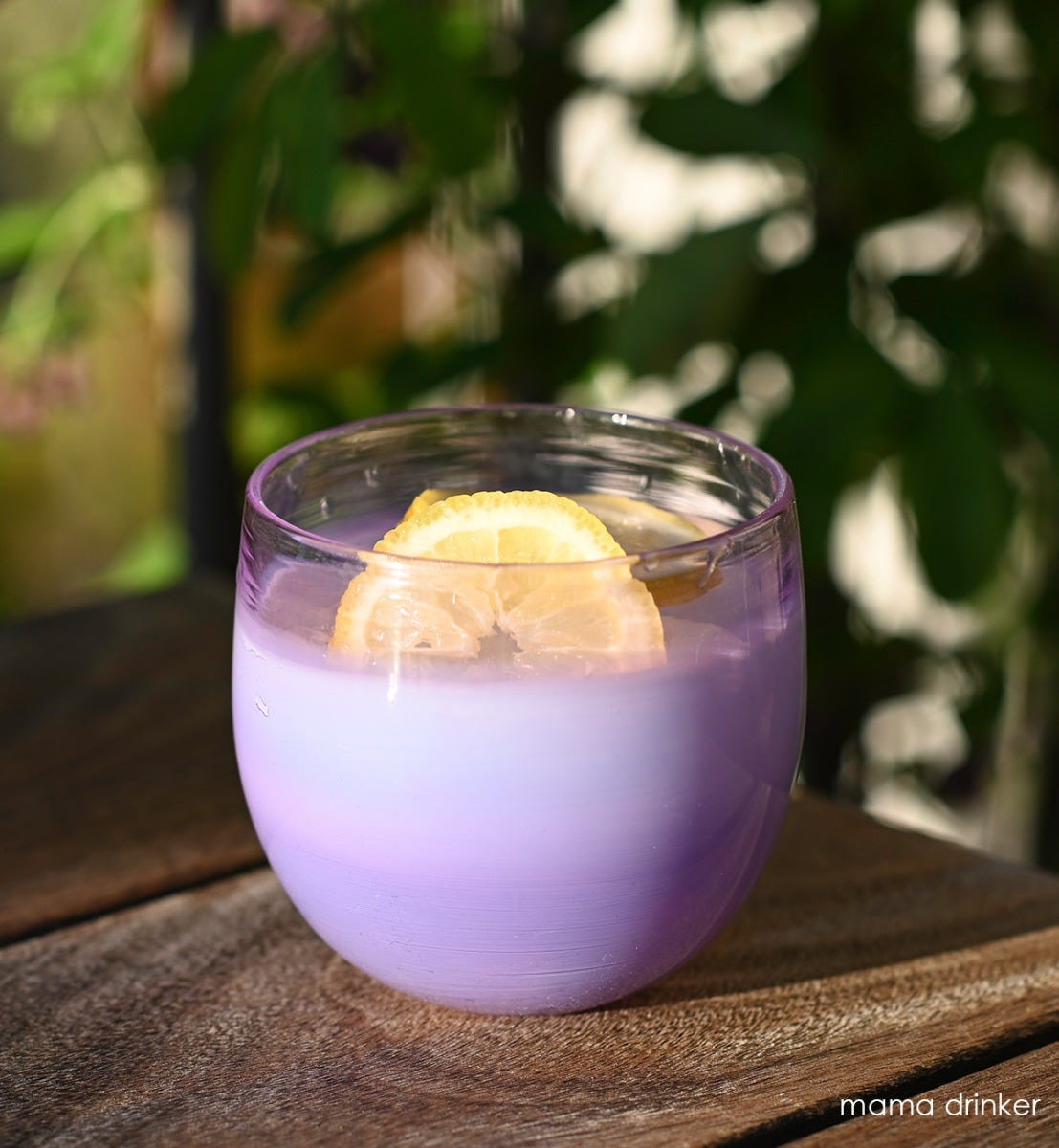 mama drinker, gradient purple, hand-blown drinking glass filled with liquid and lemons on a wood table with plants in background.