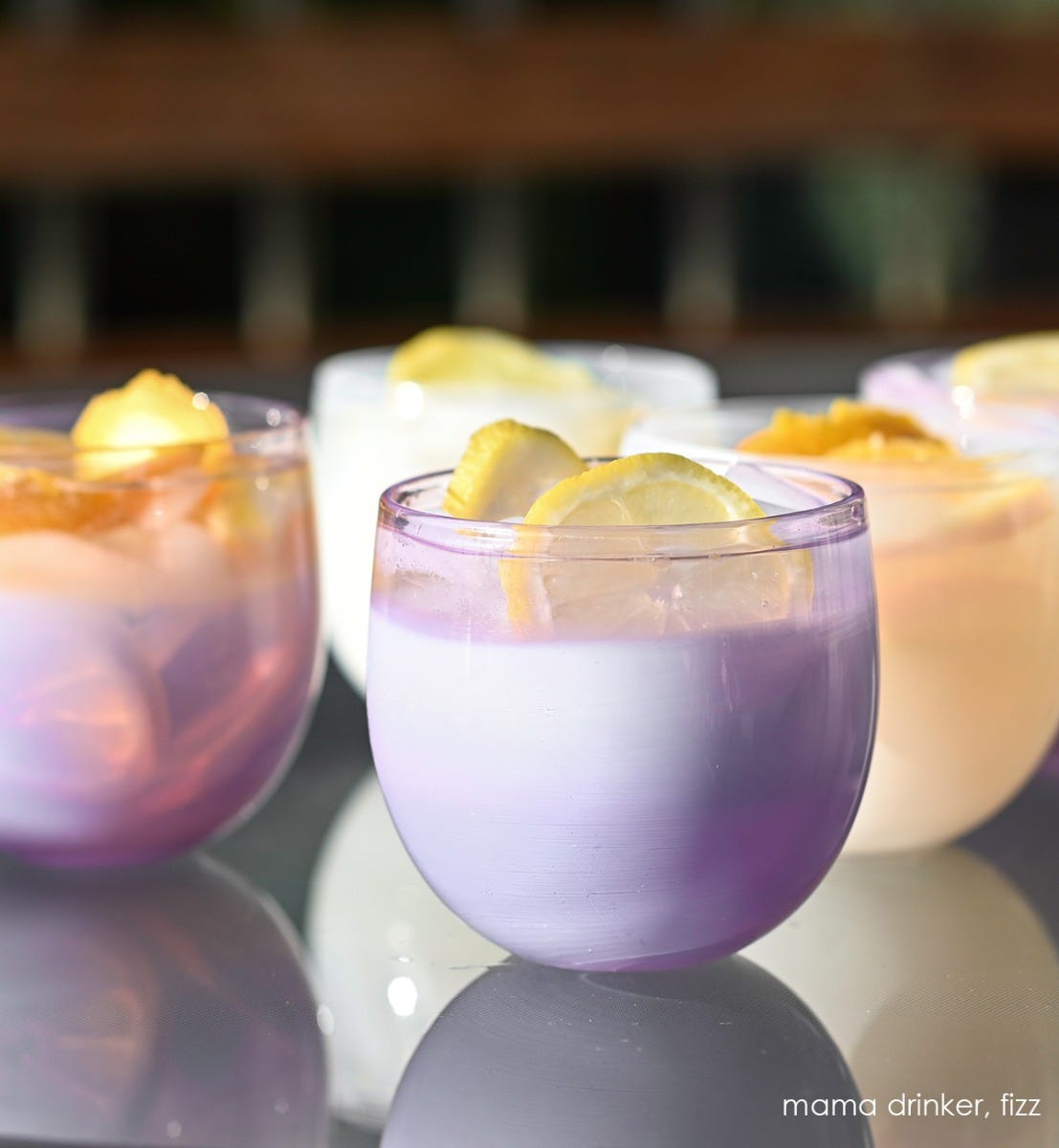 two mama drinkers, gradient purple, hand-blown drinking glasses with fizz drinkers filled with liquid and lemon on a reflective surface.