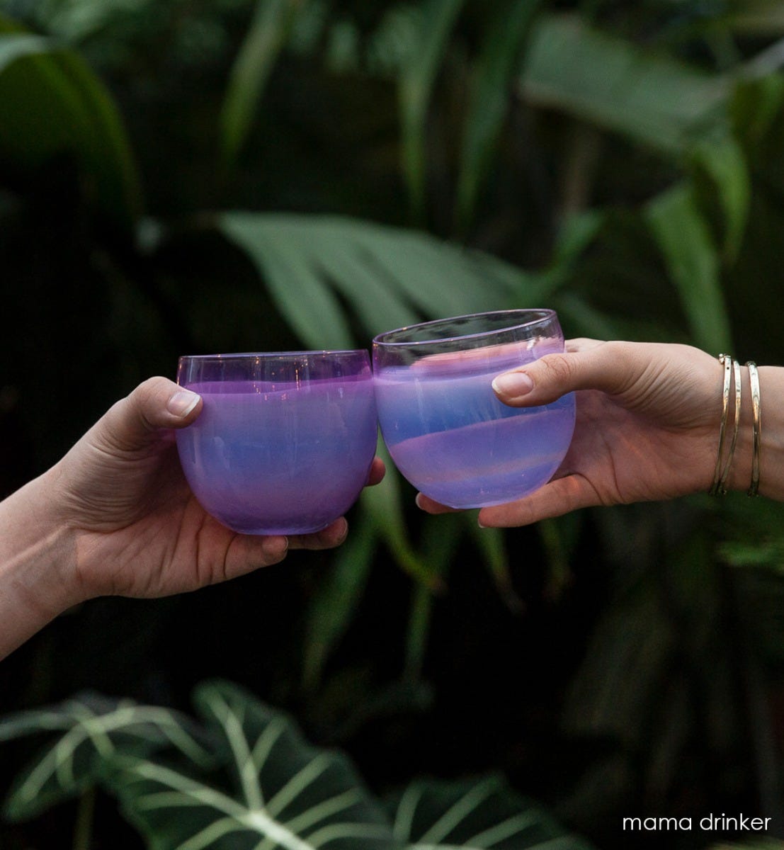 two people toasting with mama drinkers, gradient purple, hand-blown drinking glasses in front of large leafed green plants.
