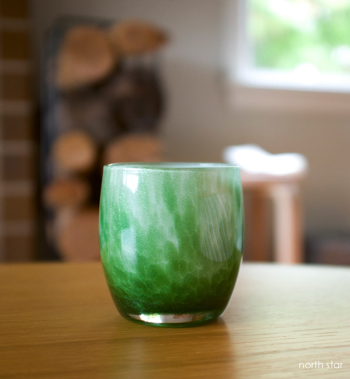 northstar, green with glitter hand-blown glass votive candle holder on a wood table with chopped firewood blurred in background.