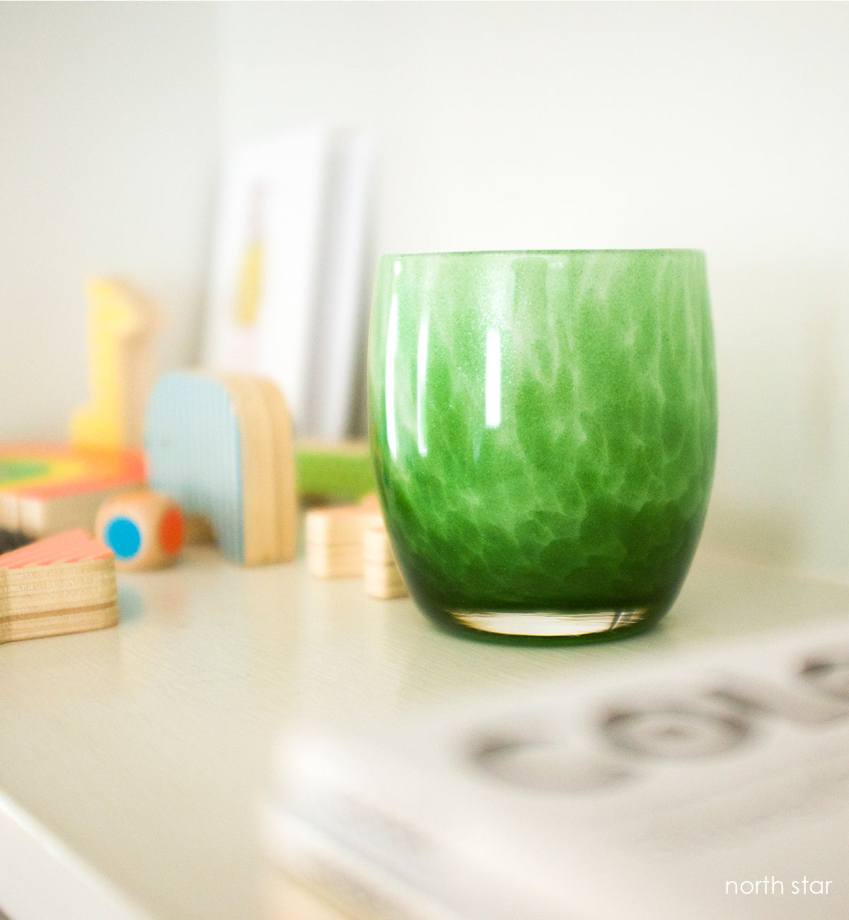 northstar, green with glitter hand-blown glass votive candle holder on a cream surface with block toys next to it.
