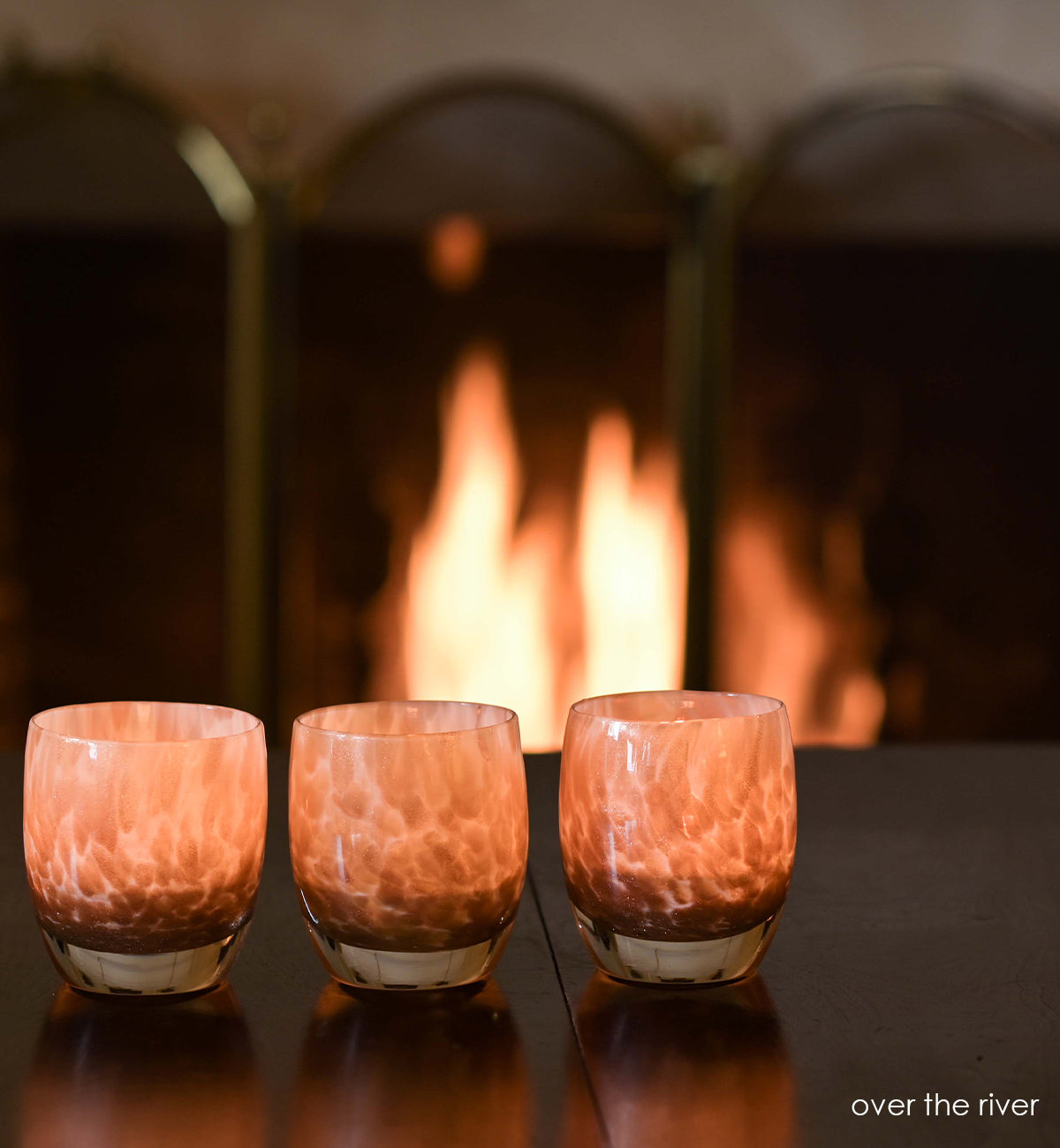 three over the river, gold glitter swirled petals, hand-blown glass votive candle holders on a wood table in front of a fire