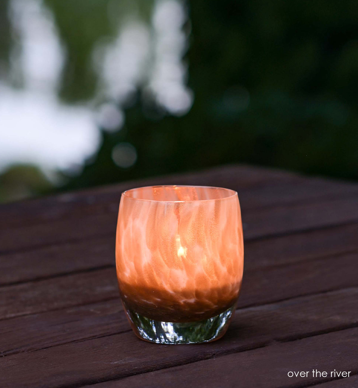 over the river, gold glitter swirled petals, hand-blown glass votive candle holder lit on an outdoor wood table.