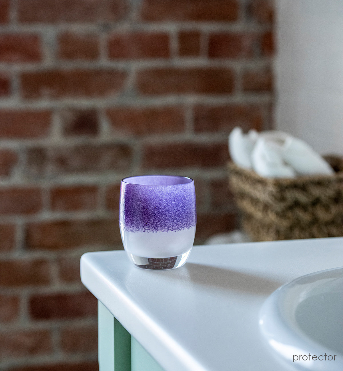 protector purple on white, hand-blown glass votive candle holder next to a bathroom sink with brick and towel basket behind.