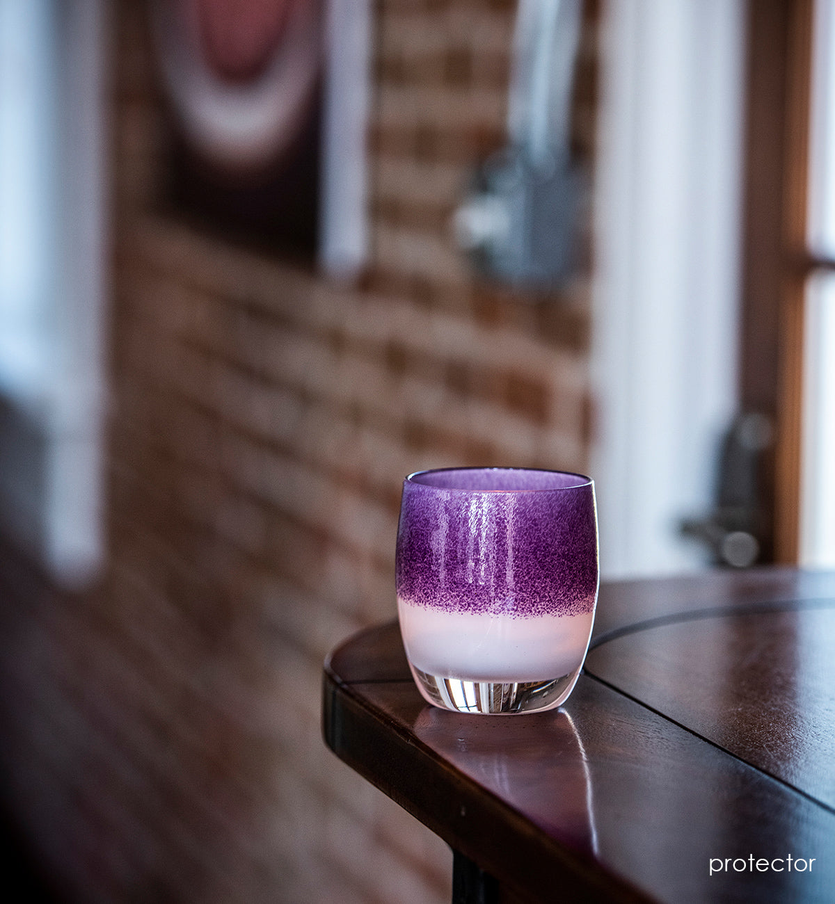 protector purple on white, hand-blown glass votive candle holder, sitting on a dark wood counter backed by exposed brick.