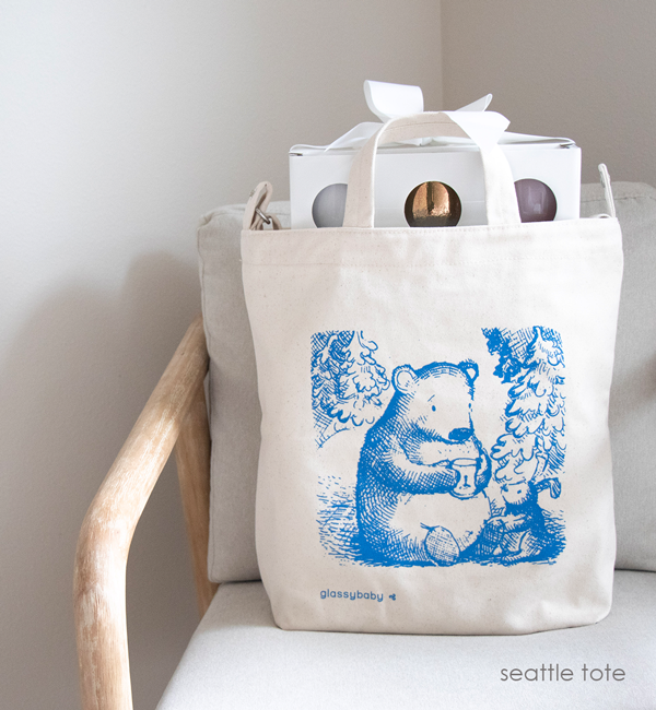 blue bear holding glassybaby, "crafted in seattle" canvas tote on a white and light wood chair with a gift set of three glassybaby peaking out.