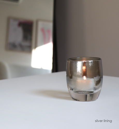 silver lining, chrome topped clear, hand-blown glass votive candle holder on a white table with mirror in background.