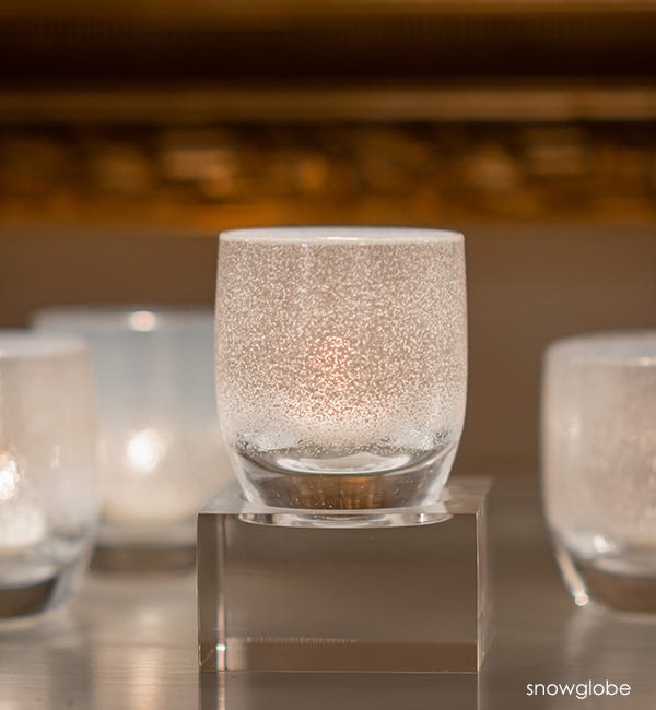  several snowglobe hand-blown white frosted glass votive candle holders, on square baby stand and gray surface with gold frame blurred in background.