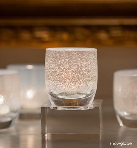 several snowglobe hand-blown white frosted glass votive candle holders, on square baby stand and gray surface with gold frame blurred in background.