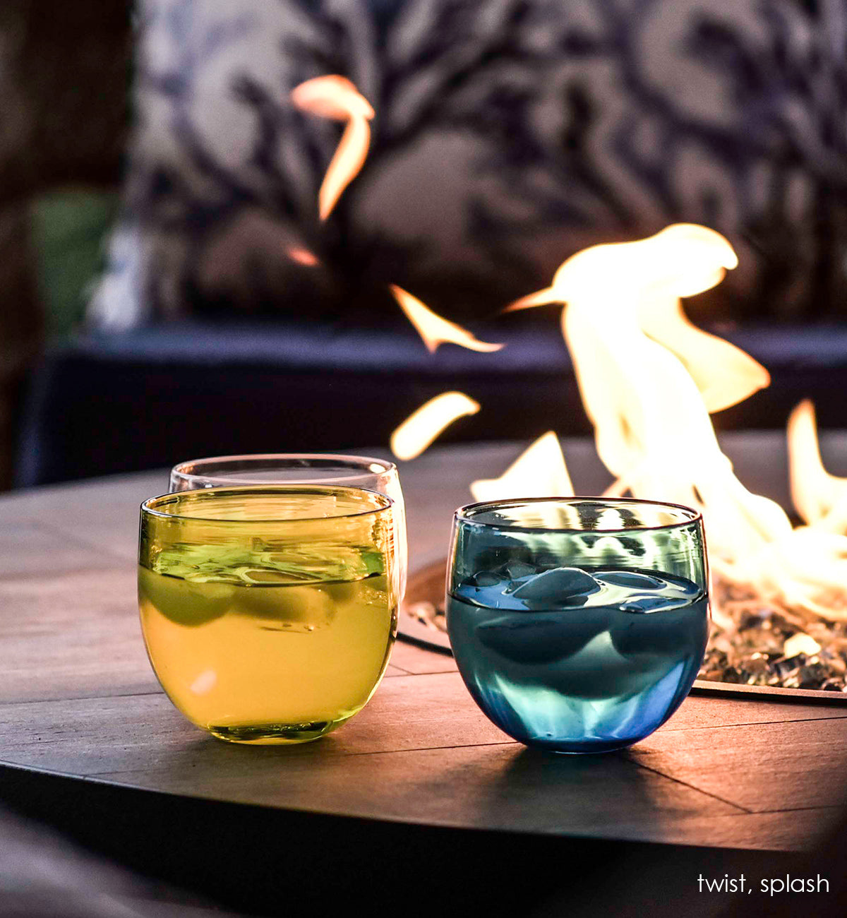 splash, hand-blown blue drinking glass with twist, both filled with ice and liquid next to an outdoor fireplace.