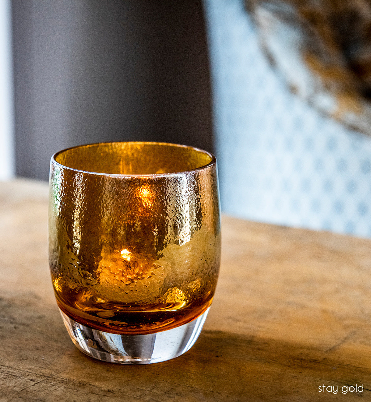 stay gold amber topped with gold, hand-blown glass votive candle holder atop a rustic wood table with blue pattern chair behind.