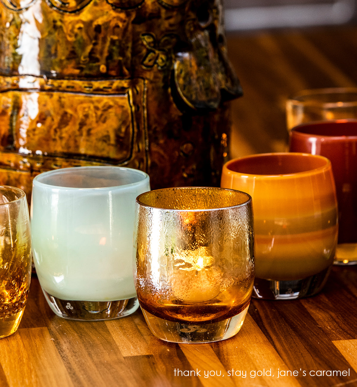 stay gold amber topped with gold, hand-blown glass votive candle holder. paired with thank you pale green, and jane's caramel on a striped wood table.