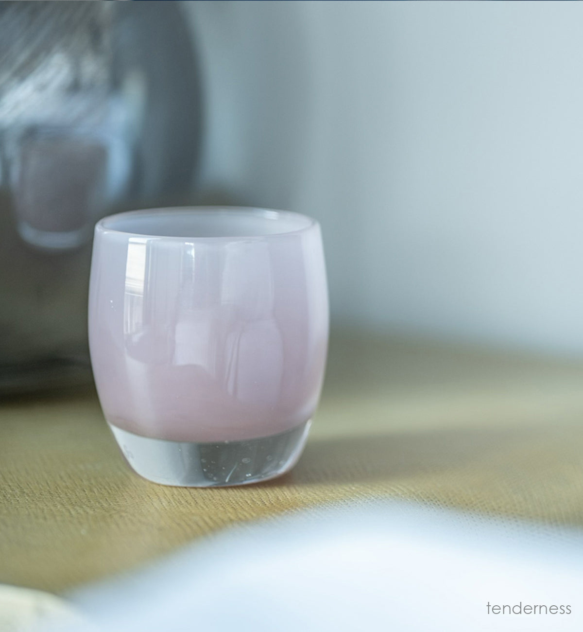 tenderness, a soft-purple hand-blown glass votive candle holder on a table with a lamp base and book.