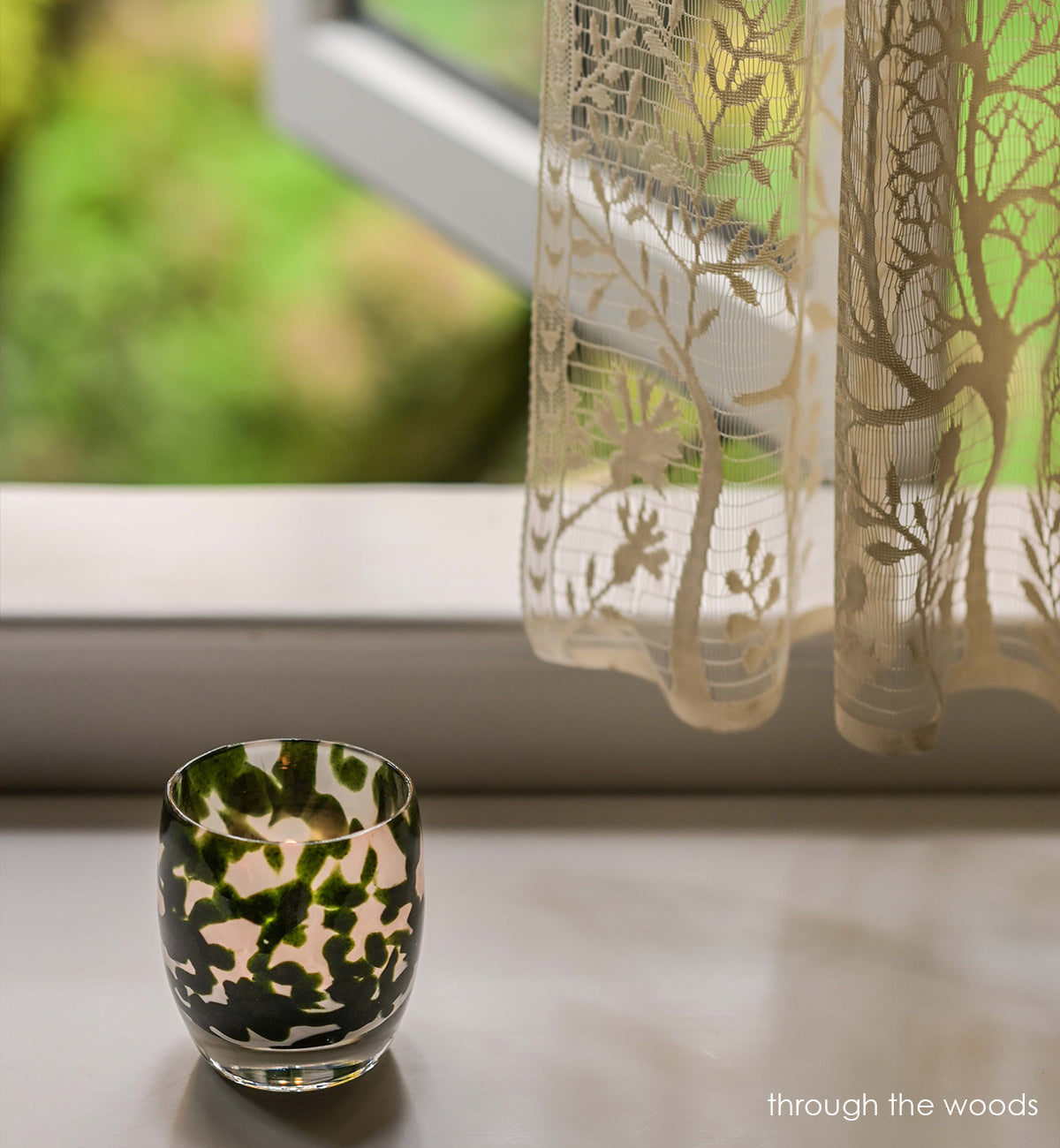 through the woods hand-blown dark green with glitter glass votive candle holder on a surface in front of an open window with lace curtains.