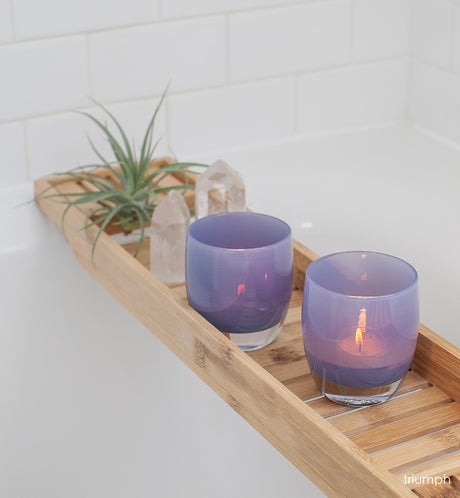 two triumph, misty purple, hand-blown glass votive candle holders on a wood bath tray next to crystals and a plant.
