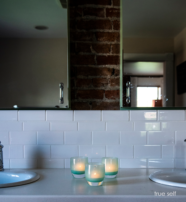 three true self, opaque translucent seafoam green, hand-blown glass votive candle holders placed in between two bathroom sinks.