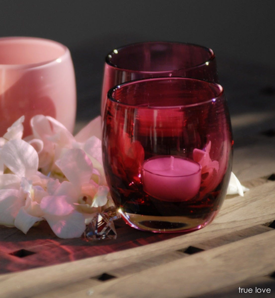 two true love, transparent deep raspberry pink, hand-blown glass votive candle holders next to white flowers.