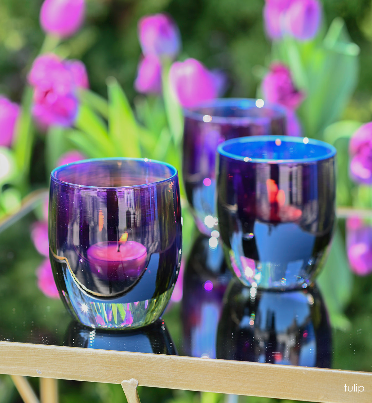 three tulip, dark purple with silver metallic interior, hand-blown glass votive candle holders on an outdoor mirror patio table with purple tulips blurred in background.