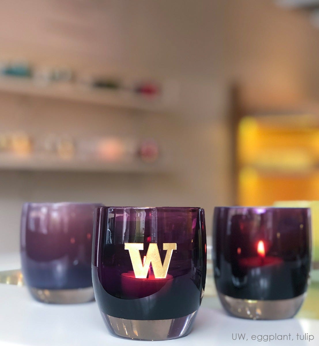 uw, purple with sandblasted university of washington etching hand painted in gold, hand-blown glass votive candle holder. Paired with eggplant and tulip on a white table in a glassybaby retail store.
